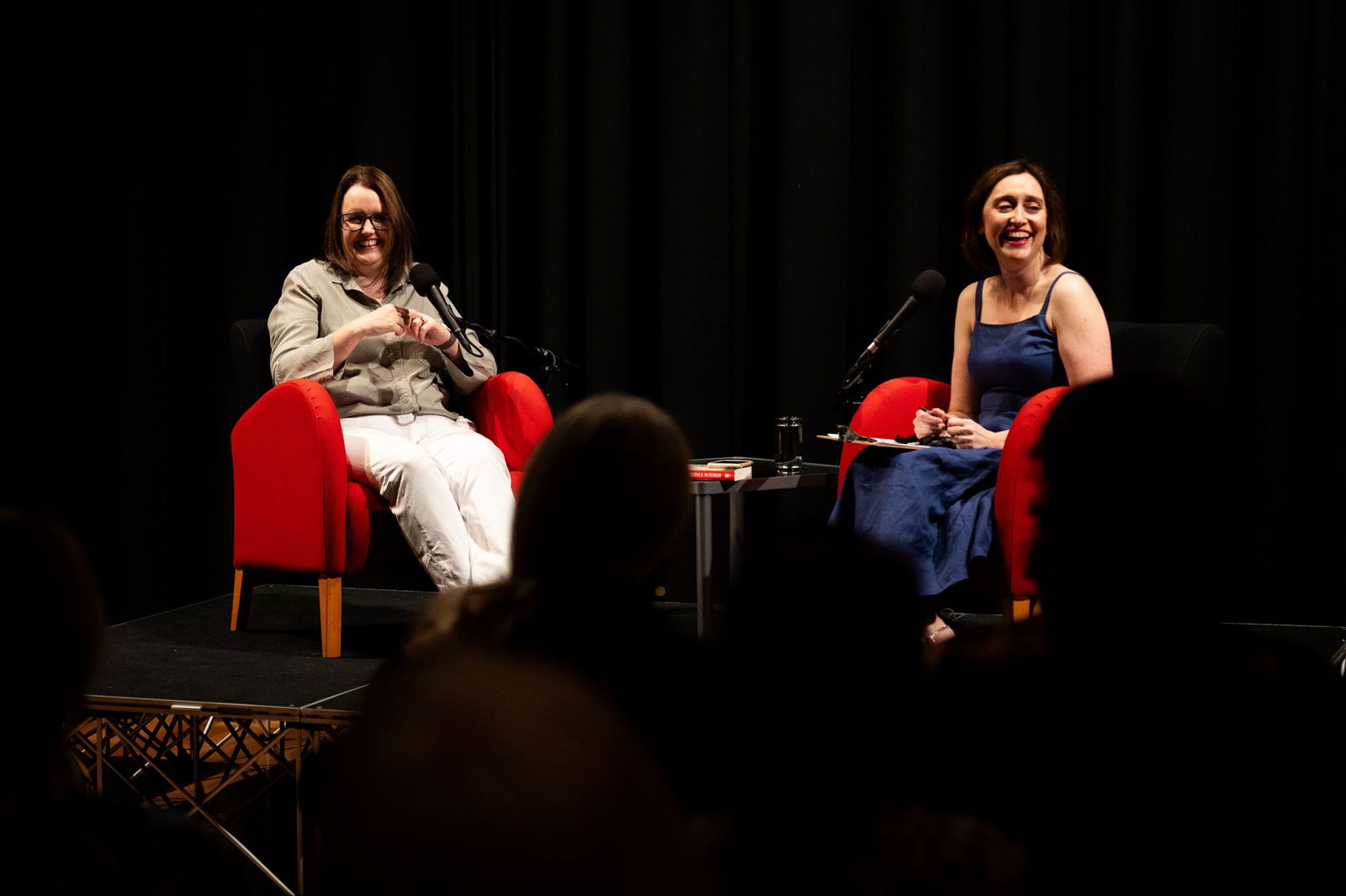 Two women laugh, sitting in red chairs side by side on a stage, with microphones in front of them.