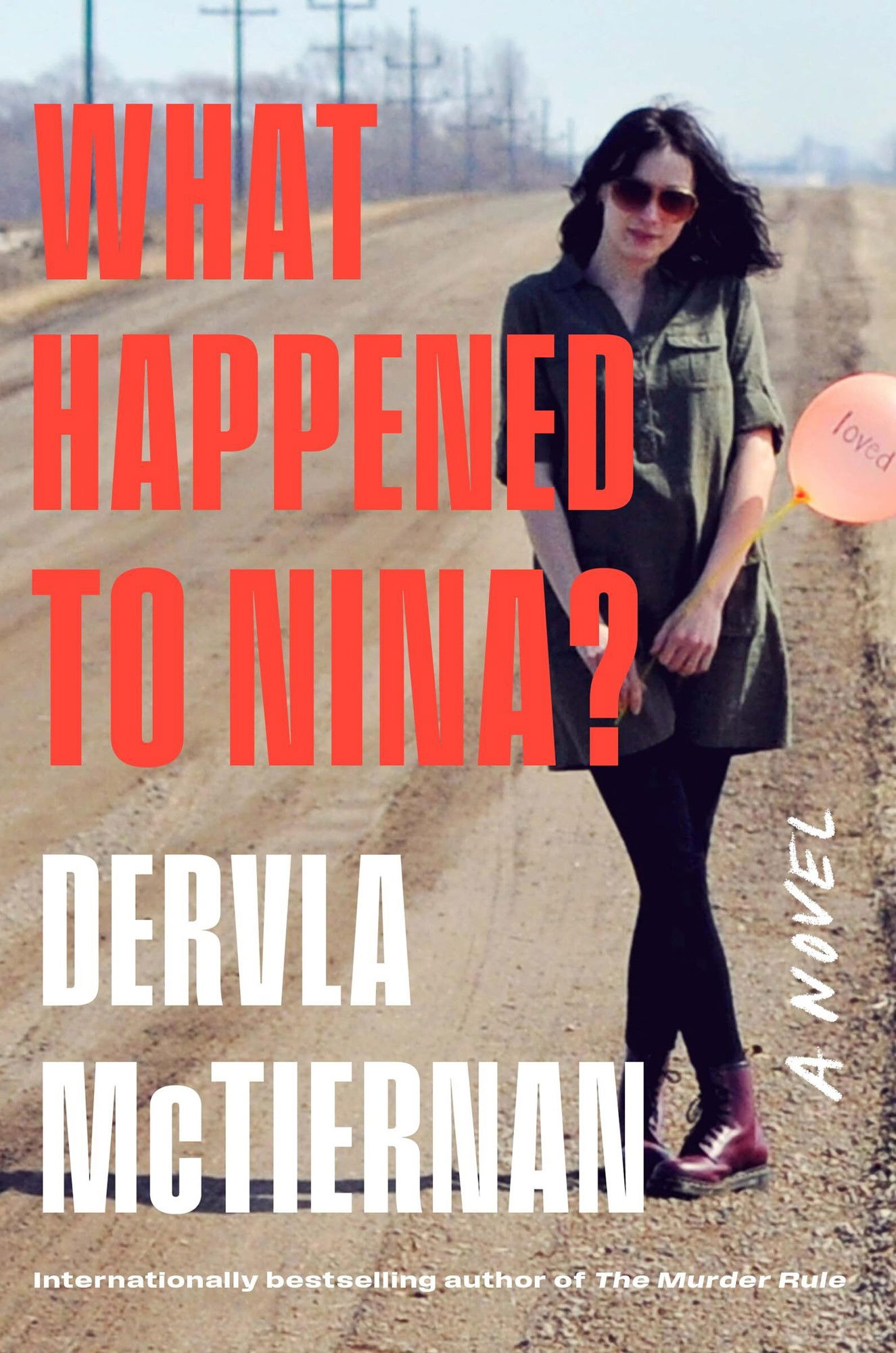 A book cover depicting a white woman with dark hair, wearing black tights, red boots and a khaki dress, standing by the side of an empty road.