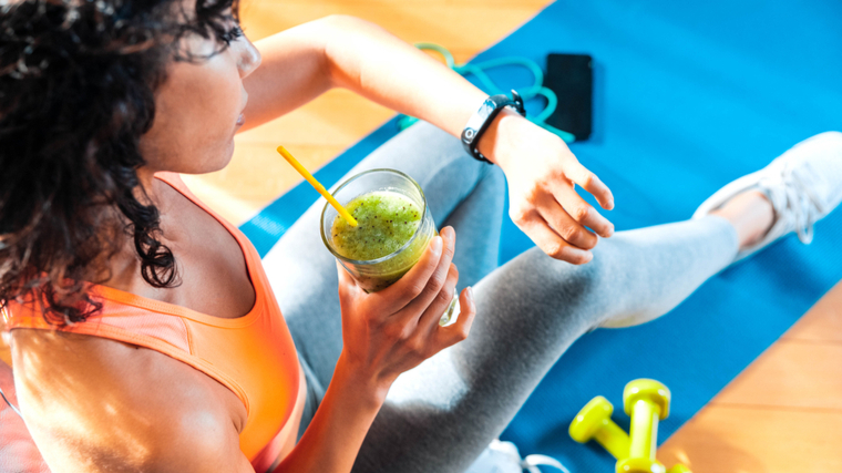 A person with curly hair wears an orange sports bra and sits on a yoga mat while holding a green smoothie and checking their watch.