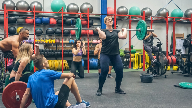 A person smiles as they perform a clean with their fellow gymgoers cheering them on.