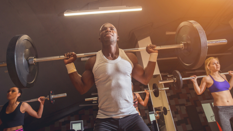 A person wearing a white tank top prepares to back squat with three people behind them, also preparing to back squat.