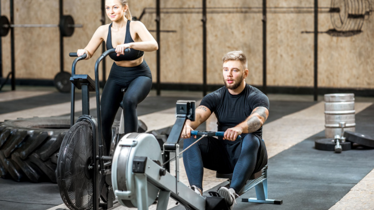 A person uses an air bike next to a person using a rower at the gym.