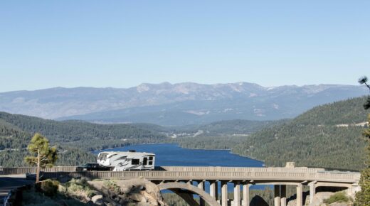 An RV driving on a scenic road; a tree-lined lake and mountains appear in the background.