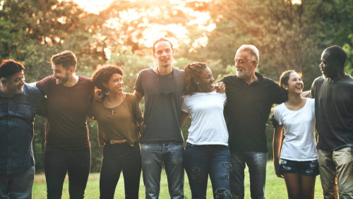 A group of people looking happy, they have their arms around each other and are in a park type setting with sunlight behind them