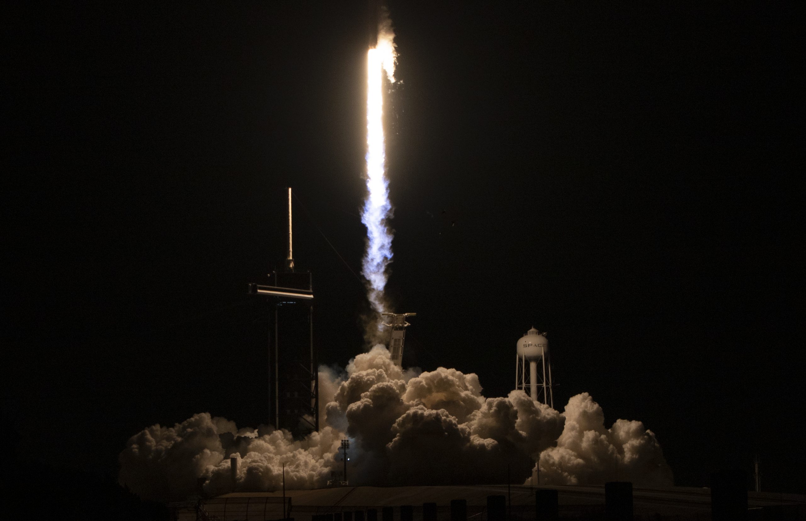 A rocket launches, trailed by bright blue and warm white flames that leave a pile of billowy smoke clouds on the ground.