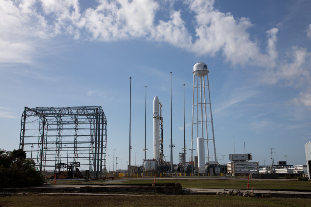 A large, white rocket with the words “ANTARES” in black letters stands vertical on its launch pad, surrounded by four poles used for lightning protection. To the right of the rocket, a water tower with long, spindly white legs has a white dome on top, with a dark logo that’s not fully visible. To the left of the rocket, is a large, open metal structure with a smaller circular structure on its side. There are several tall, gray light poles placed around the road. All of this is framed against a light blue sky with fluffy white clouds.