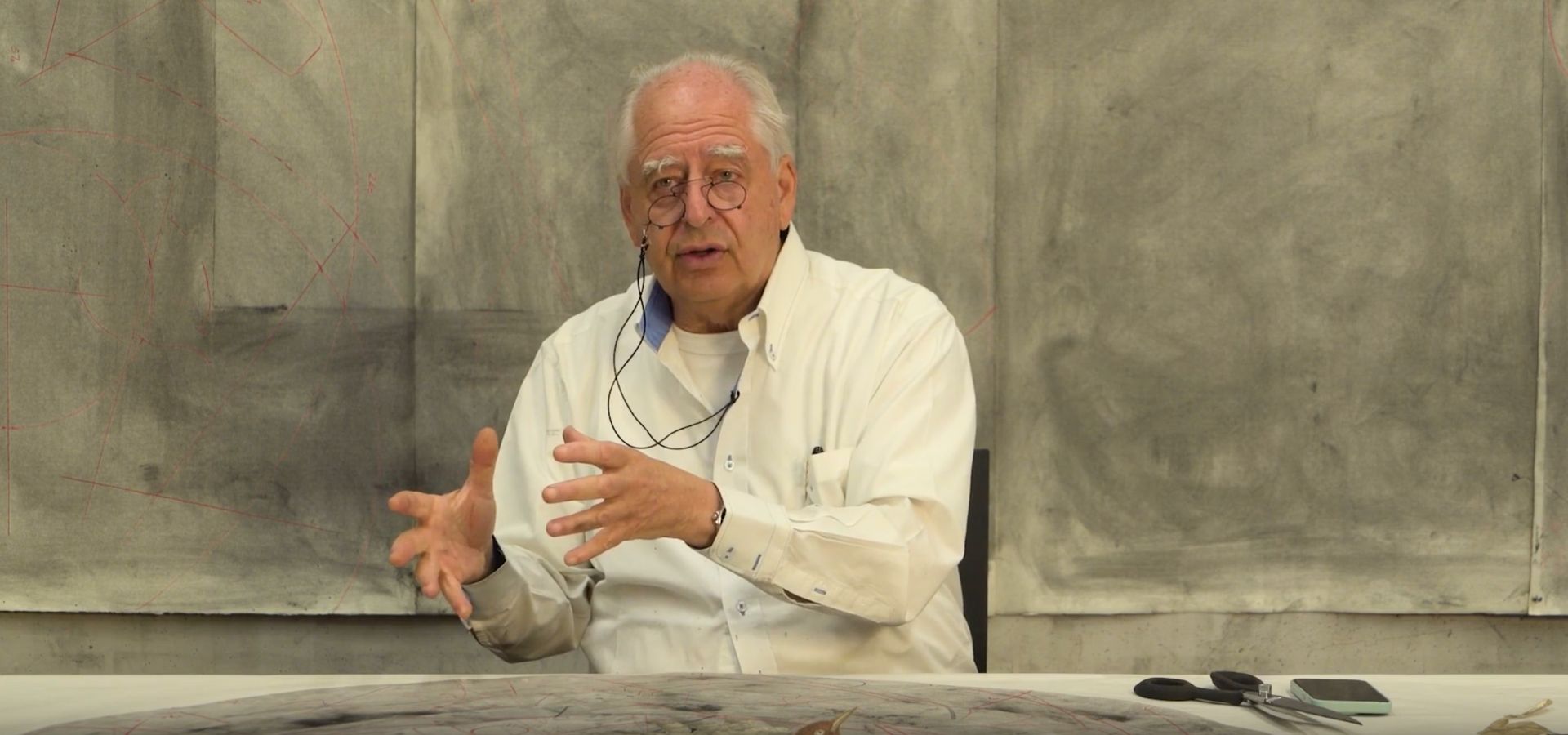 William Kentridge, an older man wearing a white dress shirt and glasses, sits at a table against a gray background and gestures with his hands.