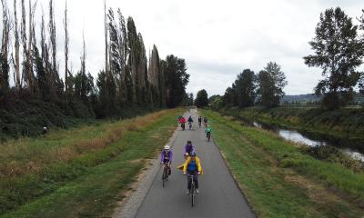 Riders zoom along a multi-use trail next to a river and a line of tall trees.