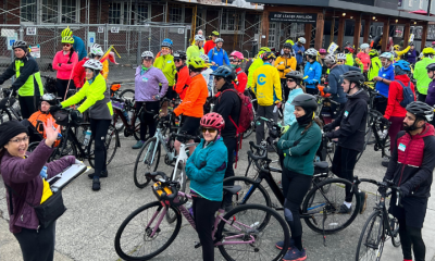 A large group of riders gathers in front of Cascade Bicycle Club. An organizer holds a clipboard and waves.