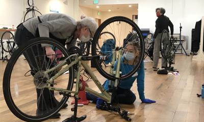 An instructor and student stand and sit behind an upside-down bike in a classroom setting.