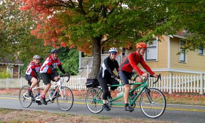 Two sets of riders on tandem bicycles pedal past beautiful fall trees