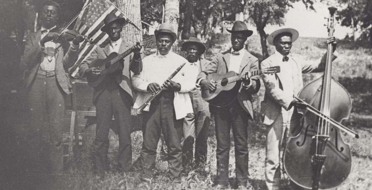Six men, the Emancipation Day Band, in the year 1900. They are carry instruments, wearing suits and hats, and in front of an American flag.