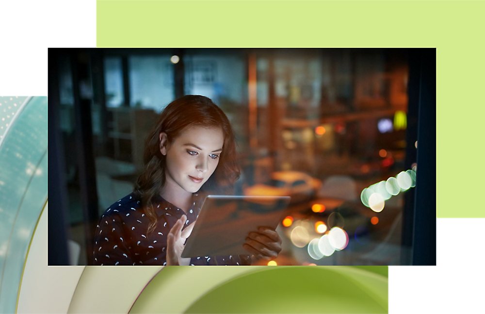 Young woman using a digital tablet at night with blurred city lights in the background through a window.