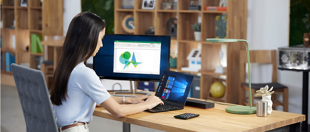 A woman works at a desk on a laptop connected to a monitor displaying graphs, in a modern office with shelving units in the background.