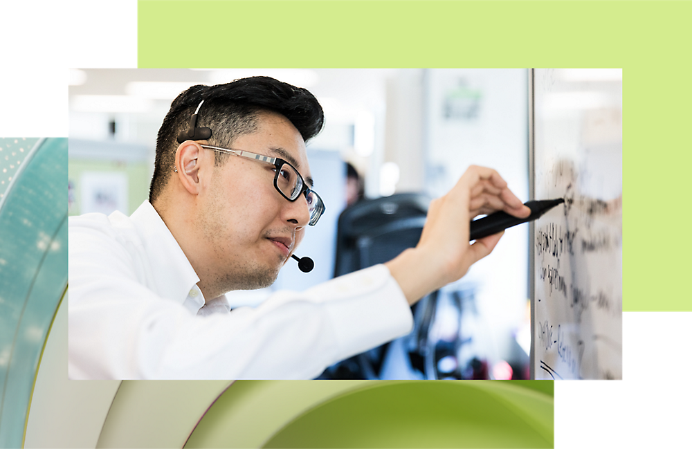 A man with glasses and a headset writes on a whiteboard in a busy office environment.