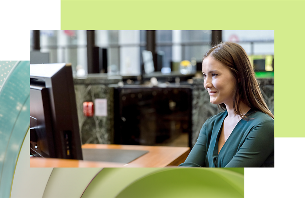 A woman in a green top smiles while using a computer in an office setting.