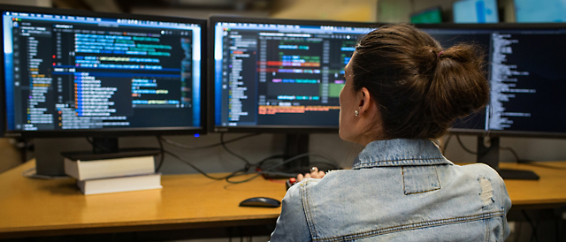 Woman coding on multiple computer screens displaying software code in a dimly lit office.