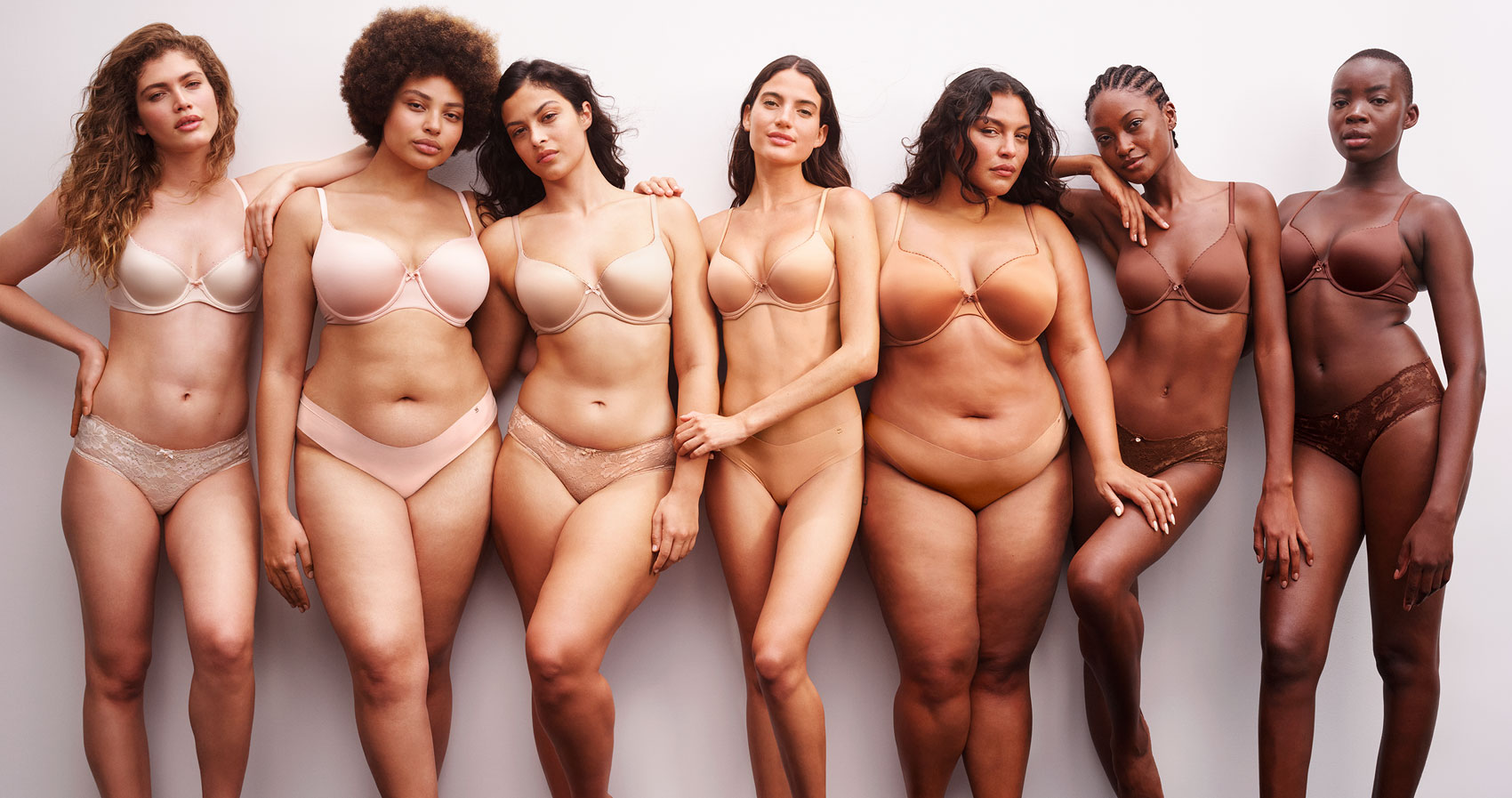 Seven VS female models of diverse body types and skin tones stand in a line, wearing matching nude-colored bras and underwear, posing against a plain white background.