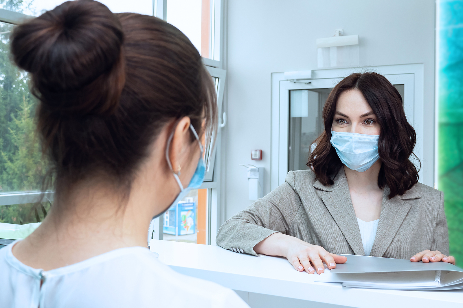 Masked health professional speaking over a desk to a masked woman with a folder