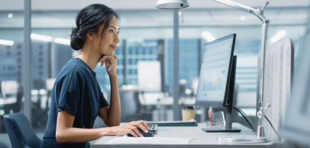 A woman in her thirties sitting at an office desk using a computer mouse and staring at a computer screen