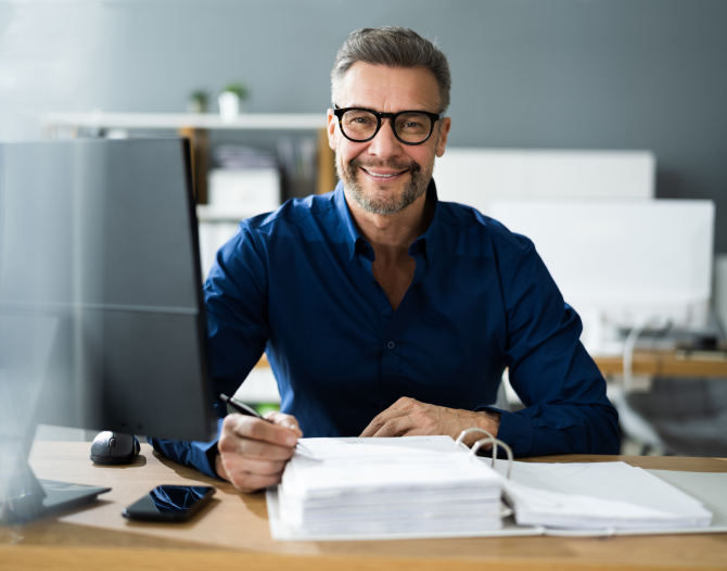 A man in his forties sitting at a desk with a computer on it, working on a stack of papers in a binder.