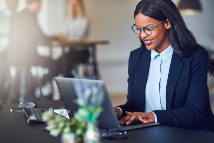 A smartly dressed woman in her thirties smiling as she works on her laptop computer.