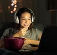 Woman smiling watching show on laptop with headphones on while eating popcorn