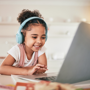 A little girl wears headphones while watching playing on her laptop. 