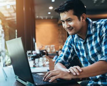 An Asian man in a blue plaid shirt types on his laptop while sitting next to a window in a coffee shop.