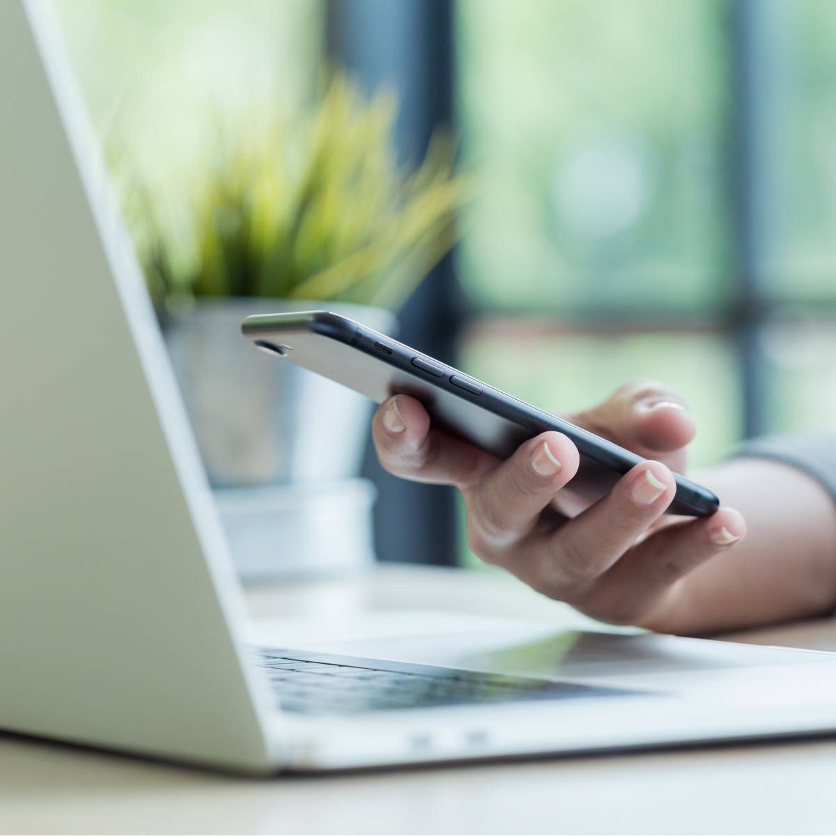 A close-up of a hand using an iPhone while hovering above a laptop.