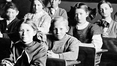 Vintage image of students at desks in a classroom with a teacher standing in the background. (education, learning)