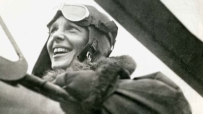 Close-up profile view of American aviator Amelia Earhart sitting in the cockpit of a helicopter. Earhart wears a bomber jacket and flight goggles on her head.