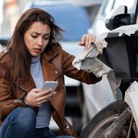 Crouching woman inspecting car accident and looking at smartphone.