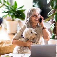 A senior woman holds her small dog while speaking on the phone.