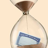 The sands of time drop on a Social Security card that sits partially buried in an hourglass.