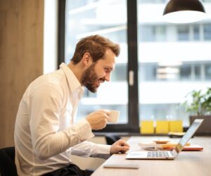 Caucasian man hunched over a corporate desk, coffee in hand, logging onto a virtual work meeting