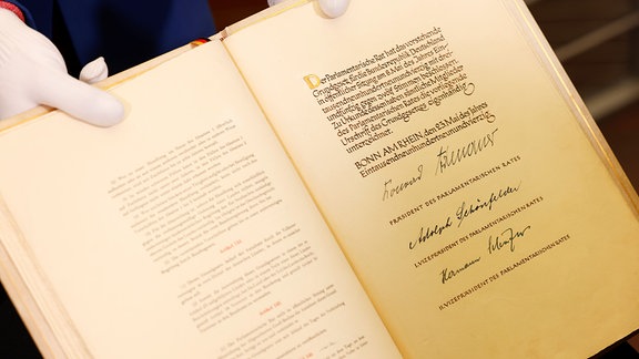 Eine Mitarbeiterin des Parlamentsarchivs des Deutschen Bundestages zeigt die Unterschrift des damaligen Präsidenten des Parlamentarischen Rates und späteren Bundeskanzlers Konrad Adenauer (M. r) und seiner Stellvertreter auf einer Seite im Originalbuch des Grundgesetzes, der Verfassung der Bundesrepublik Deutschland, während einer Pressepräsentation im Vorfeld des 75-jährigen Bestehens der Verfassung.