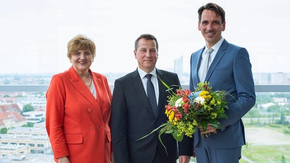 Eine Frau mit roter Jacke sowie zwei Männer in Anzügen blicken in die Kamera. Ein Mann hält einen Blumenstrauß.