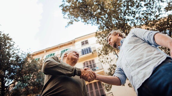 Symbolbild, von unten gesehen: Ein älterer und ein jüngerer Mann geben sich die Hand.