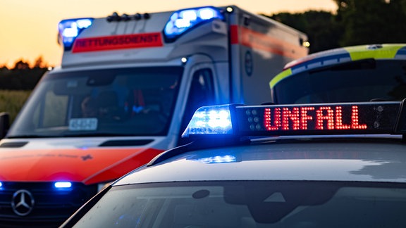 Ein Einsatzfahrzeug, Streifenwagen, der Polizei steht mit Blaulicht und dem Schriftzug Unfall im Display an einem Unfallort. Neben dem Polizeiwagen steht ein Rettungswagen, RTW.