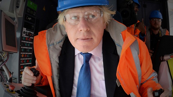 Boris Johnson mit Warnweste, Helm und Schutzbrille in einer Steuerkabine mit Joysticks, frontal, weitwinklig, geduckt und lustiger Blick zur Seite.