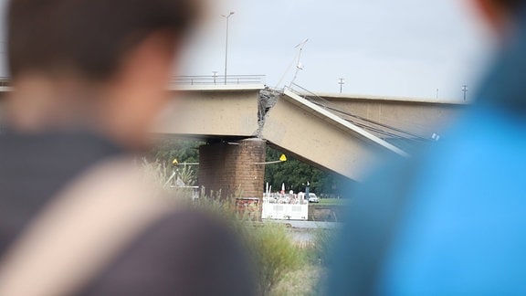 Herangezoomte abgeknickte Stelle einer Brücke, links uns rechts davon unscharf im Vordergrund Menschen