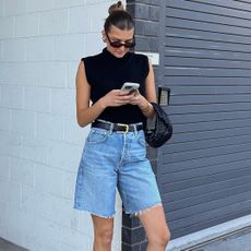 fashion influencer Sara Walker looks at her Iphone cell phone in front of a white cinder brick wall wearing a slicked back bun, black oval sunglasses, a mock neck black sleeveless top, a mini black Bottega Veneta bag black belt, and Bermuda denim cutoff shorts