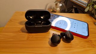 Black Sony WF-1000XM4 wireless earbuds on a wooden desk with their carry case and a phone running the Sony Headphones Connect app on screen.