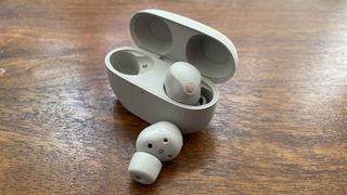 White Sony WF-1000XM5 earbuds and their charging case on a wooden surface. One earbud is inside the case, one in front of it.