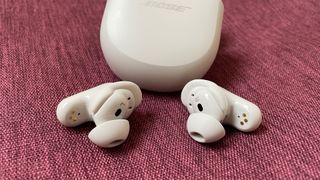 White Bose QuietComfort Ultra Earbuds lying angled outwards in front of their charging case on a red material surface.
