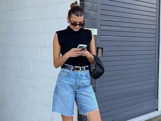 Fashion influencer Sara Walker stands in front of a white brick wall wearing a slicked-back bun, black oval sunglasses, a mock neck black sleeveless top, a mini black Bottega Veneta bag black belt, and Bermuda denim cutoff shorts.