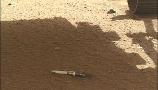 On a flat, slightly rocky ground, there is a sample tube laying there. A rover's wheel is seen toward the top right.