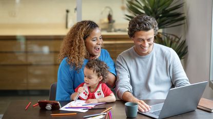 A young husband and wife smile and laugh as they look at a laptop at the kitchen table while their toddler is in the woman&#039;s lap.