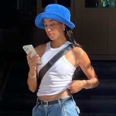 Woman wearing white tank top, blue bucket hat, and wide leg jeans shopping on phone.